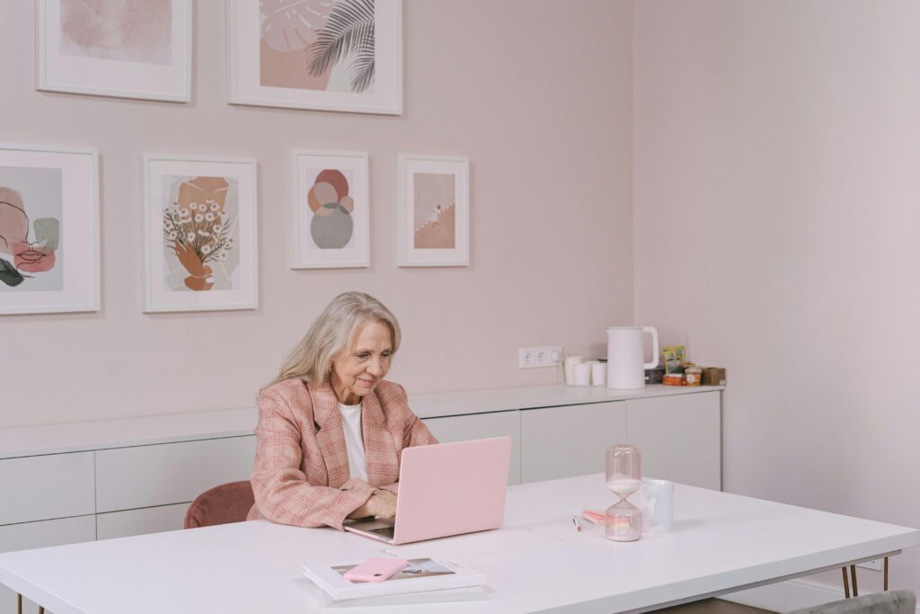 pexels-photo-7394565-7394565 Elderly woman in pink blazer uses laptop in modern home office setting, smiling and engaged.